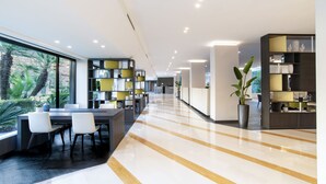 A modern hotel lobby with a long reception desk, seating area, and a large window with a view of greenery. - Mercure Eur Roma West (Rome)