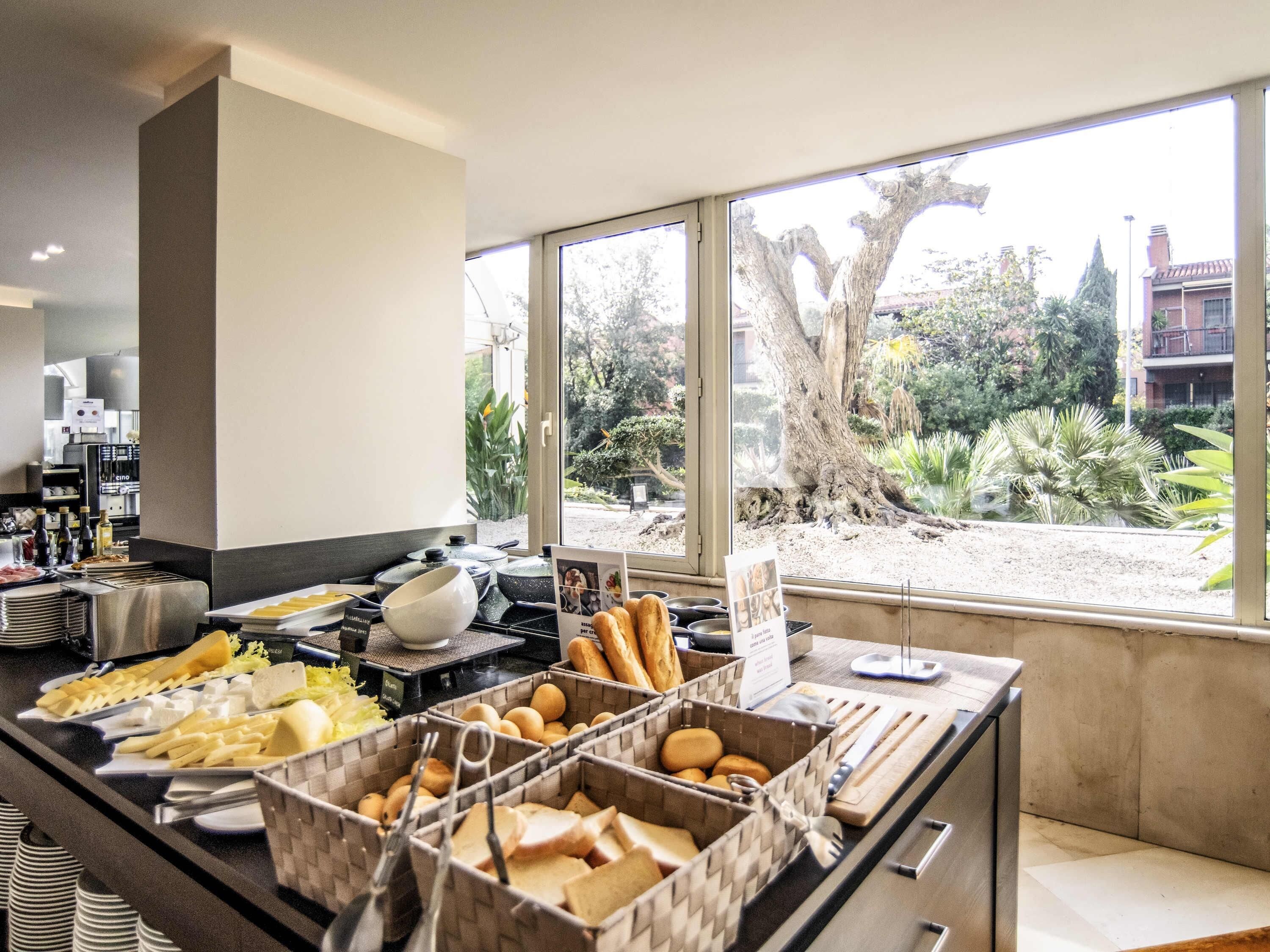 Ein Buffet mit verschiedenen Speisen und Besteck, ein Fenster mit Blick auf Bäume und Gebäude, sowie ein Küchenbereich im Hintergrund.