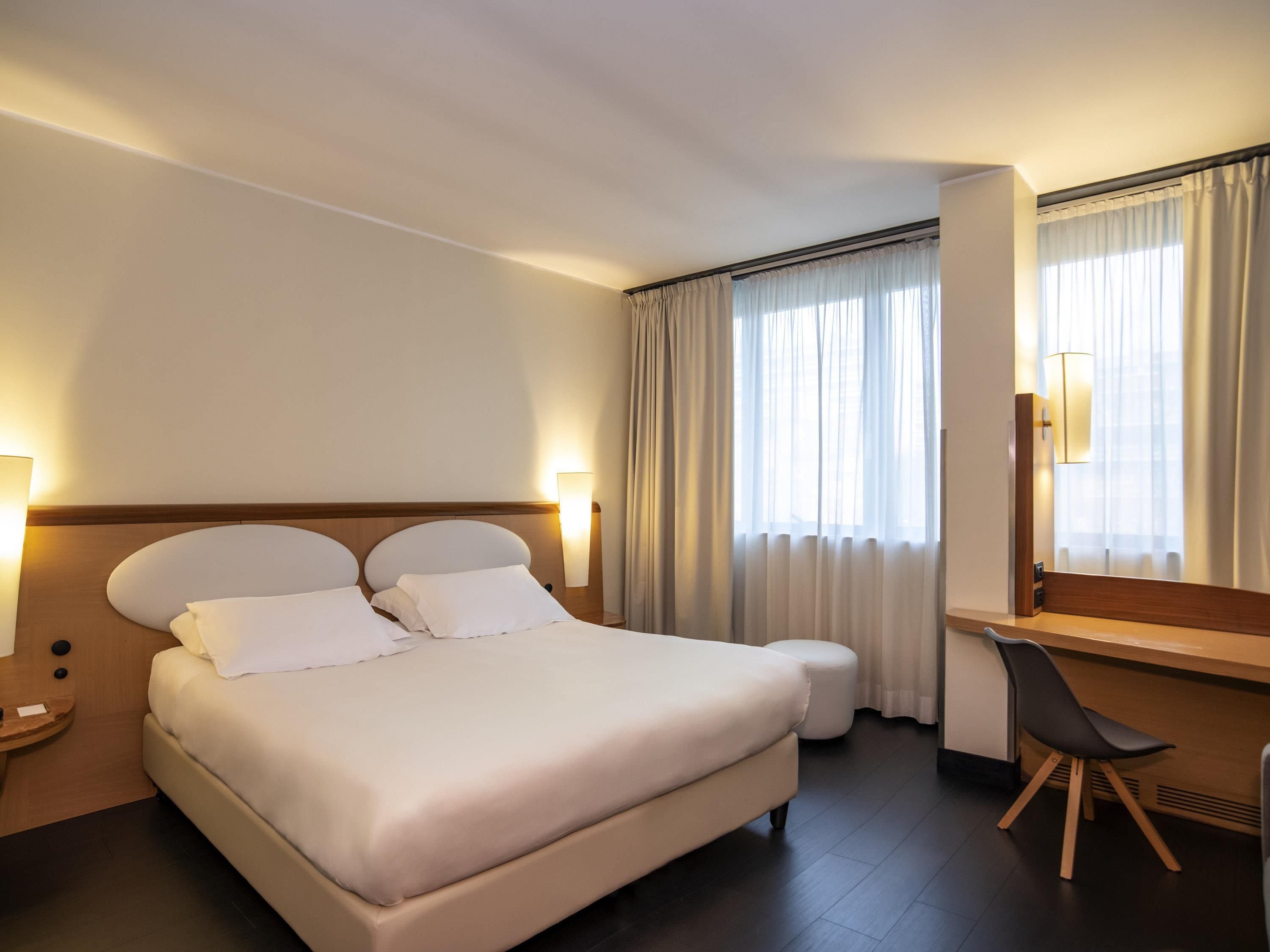 Une chambre d’hôtel avec un grand lit, des lampes de chevet, un bureau avec une chaise et une fenêtre avec des rideaux.