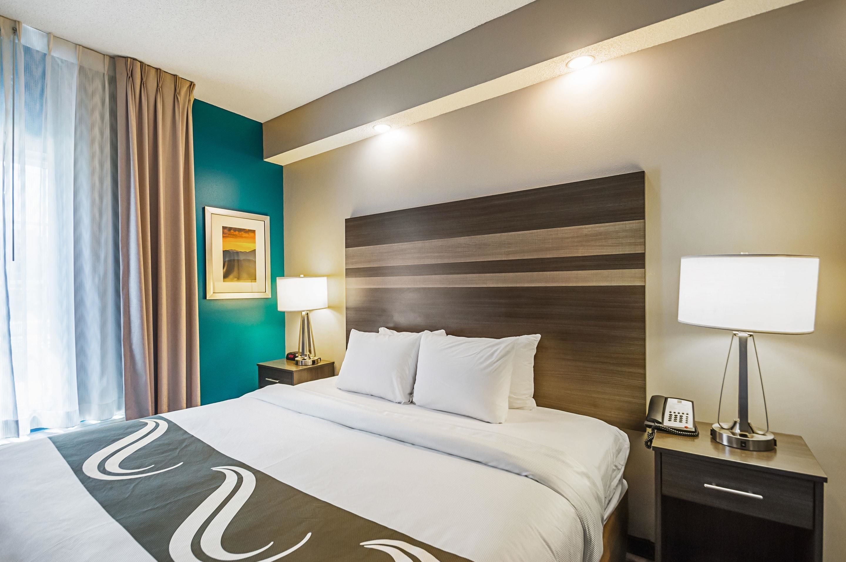 Habitación de hotel con una cama grande, dos mesitas de noche, una mesilla con lámpara y un cuadro en la pared.