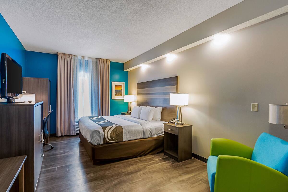 A hotel room with a bed, bedside tables, a TV, a desk, and a green chair.