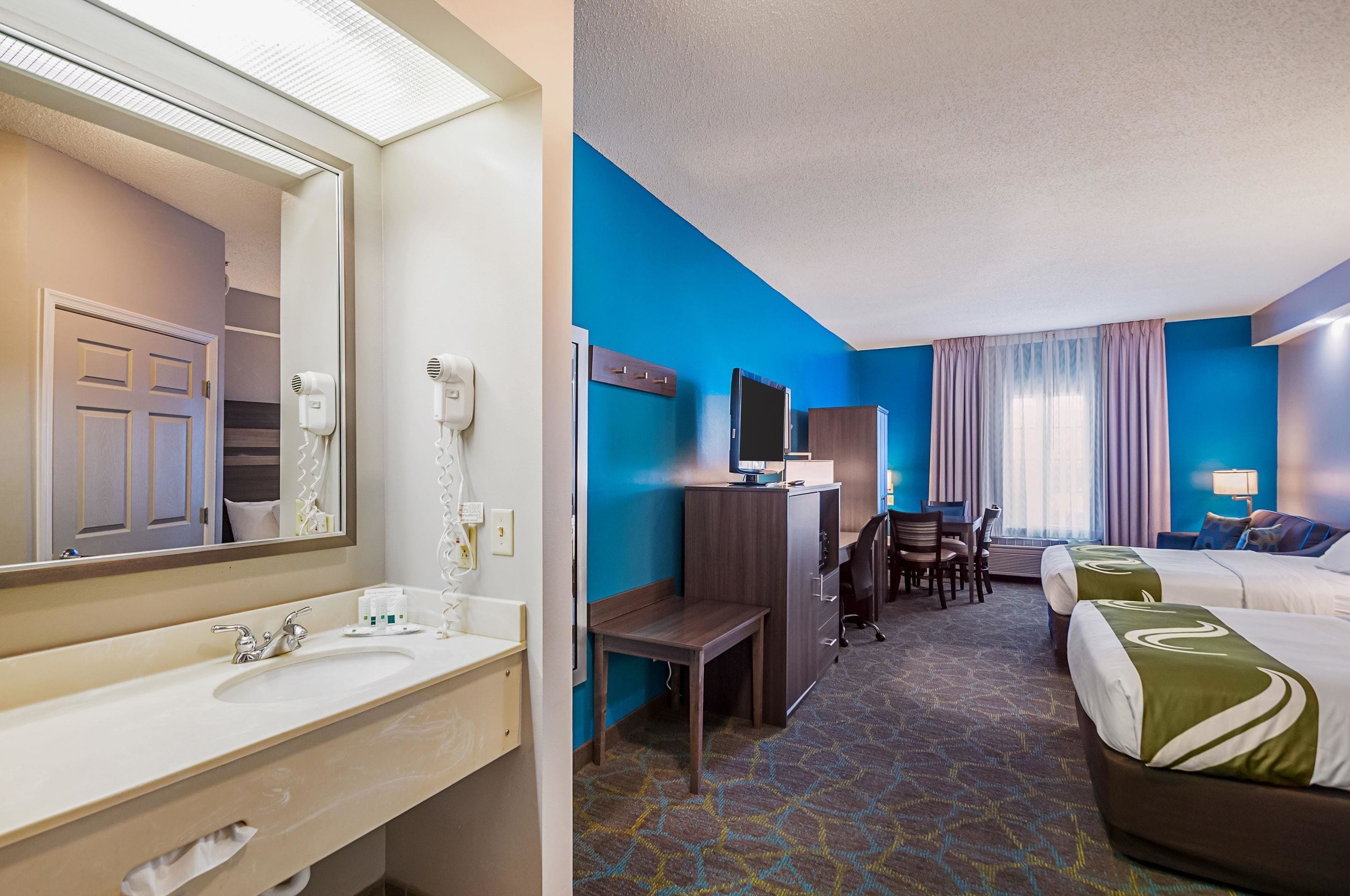 Une chambre d’hôtel avec un grand miroir, un lavabo, un bureau avec une chaise, une télévision et deux lits.
