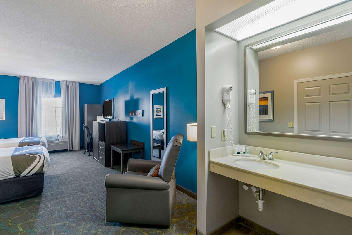 A hotel room with a bed, desk, chair, and a bathroom with a sink and mirror.