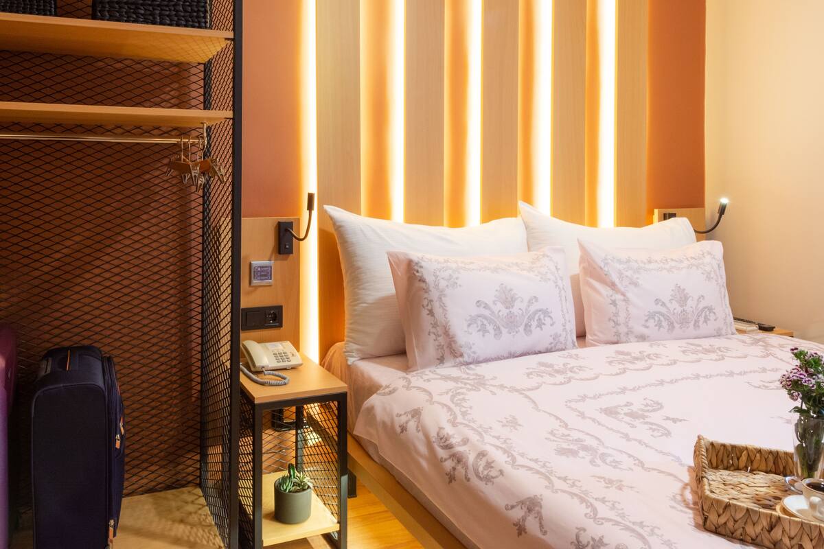 A hotel room with a bed, a bedside table with a phone, a suitcase, and a wall with vertical light panels.
