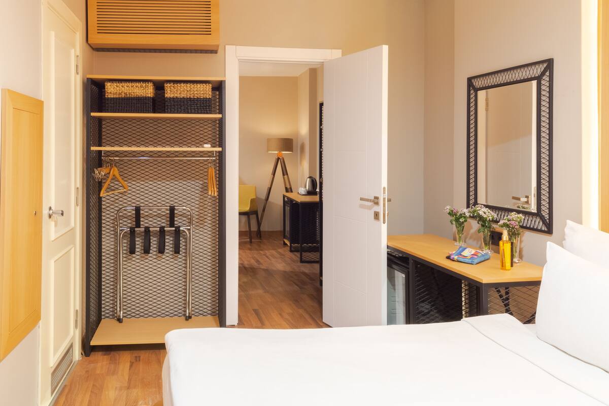 A hotel room with a bed, a wooden wardrobe, a mirror, and a desk with a lamp and flowers.