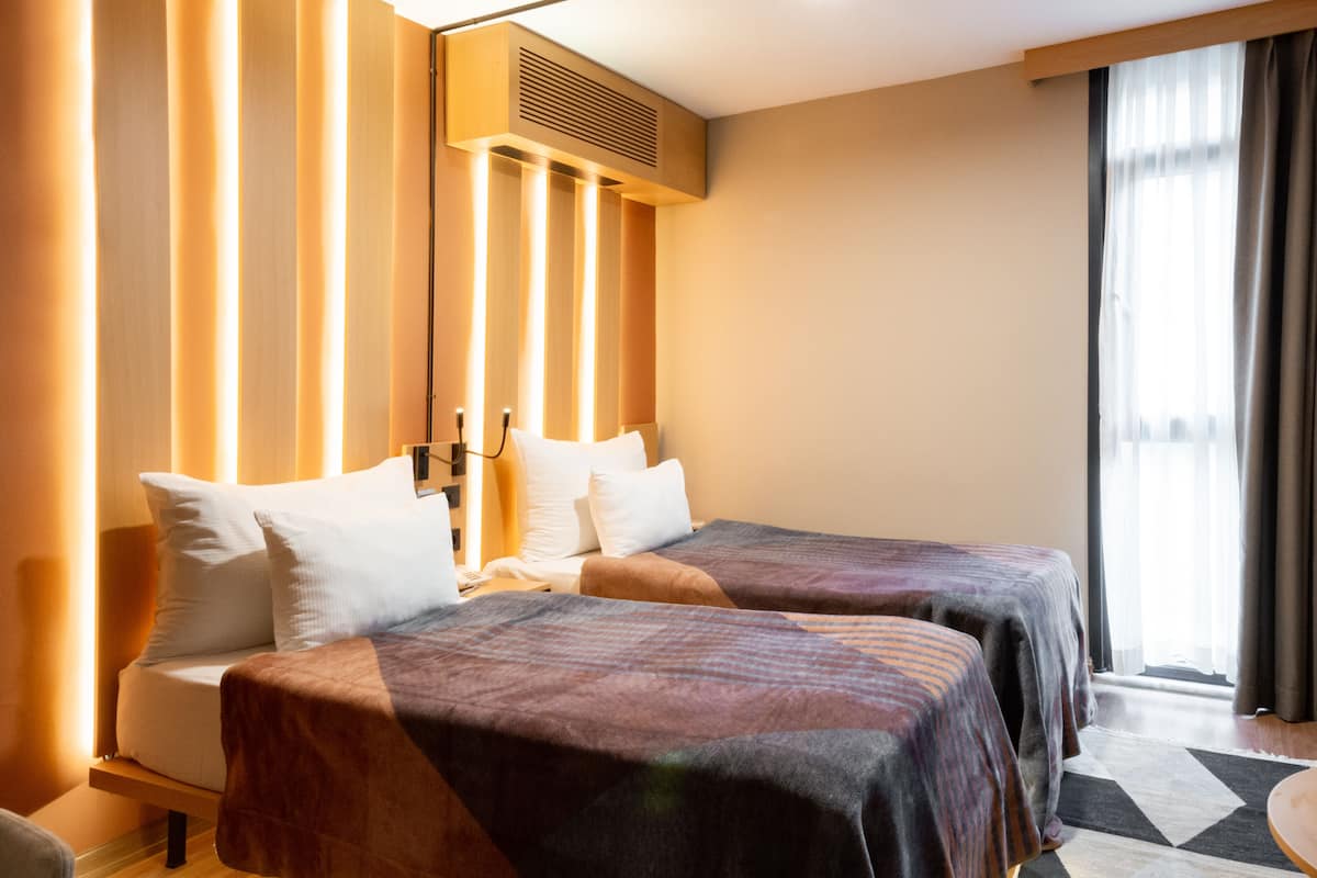 A hotel room with two beds, a large window with curtains, a ceiling light, and a wall-mounted air conditioning unit.