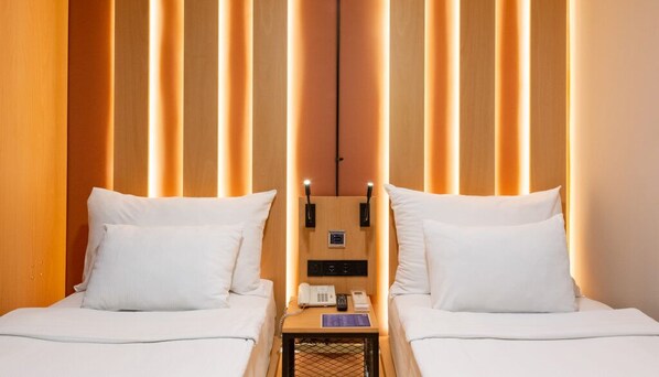 A hotel room with two beds, a small table with a telephone and a notepad, and a wooden panel wall with vertical light strips.