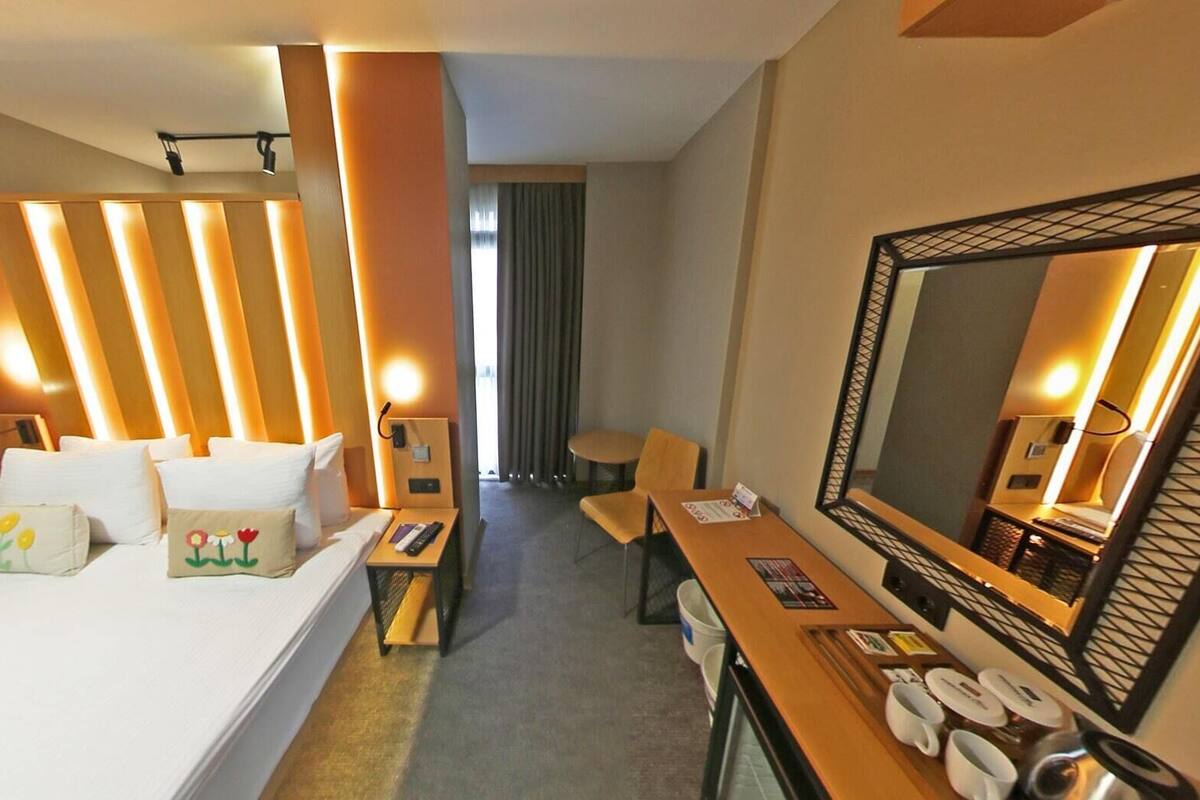 A hotel room with a large bed, a desk, a chair, a mirror, and a view of the outside.