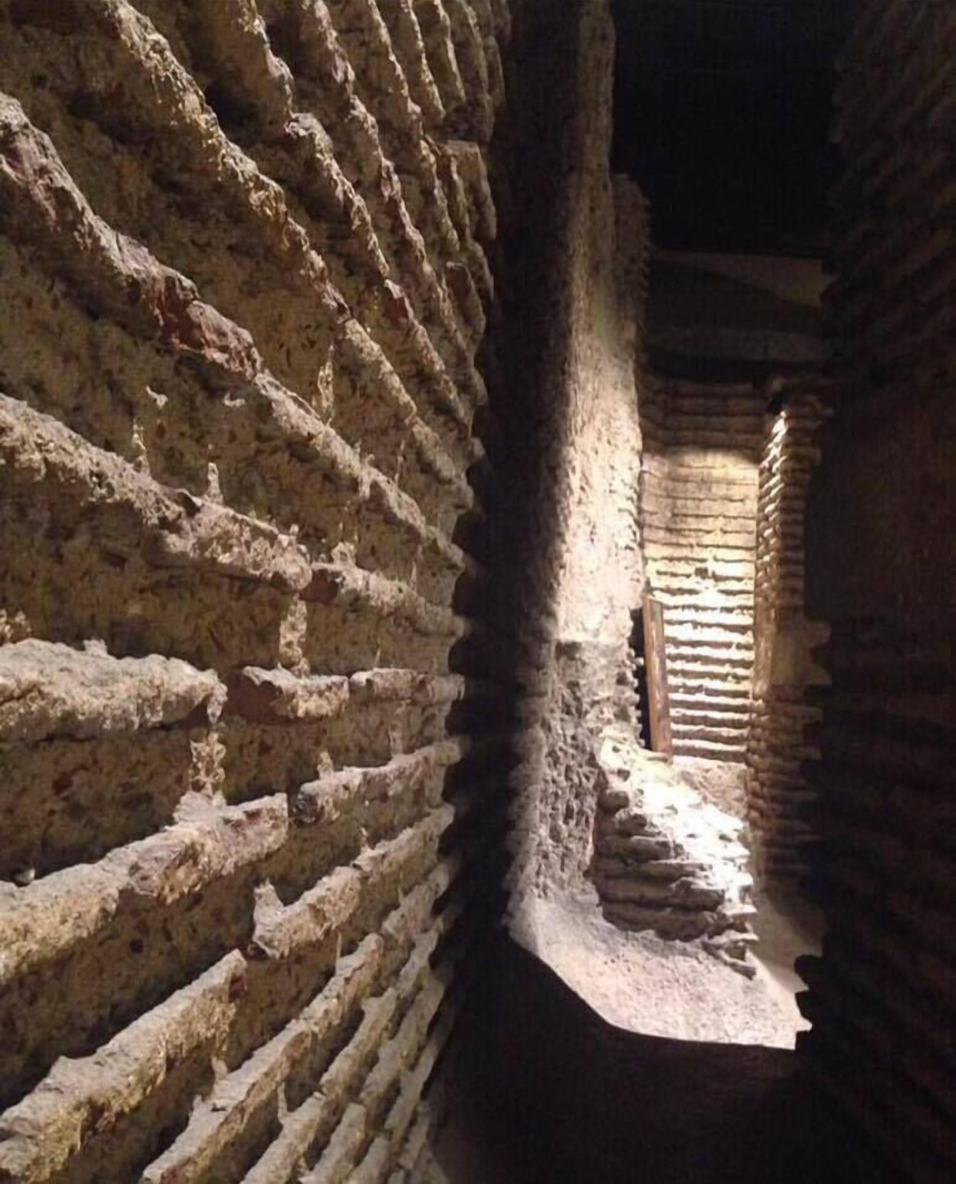 Un étroit passage aux murs de pierre brute, avec une construction en briques visible au loin.