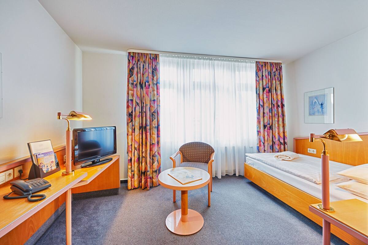 A hotel room with a bed, a desk with a computer, a chair, and a small table.