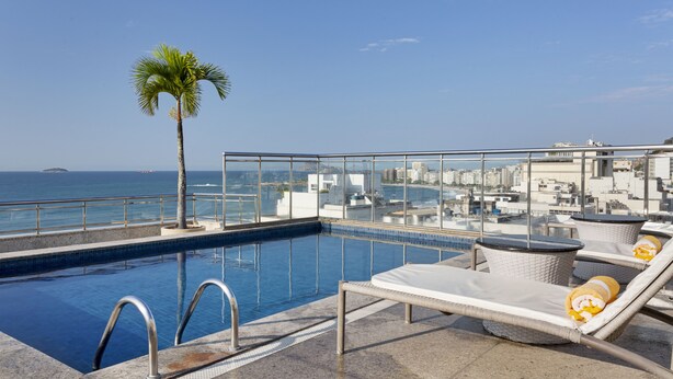 Una piscina en la azotea con sillones de descanso y una palmera, con vistas a la ciudad y al mar.