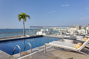 A rooftop pool with lounge chairs and a palm tree, overlooking a cityscape and the sea. - Windsor Palace Copacabana (Rio de Janeiro)