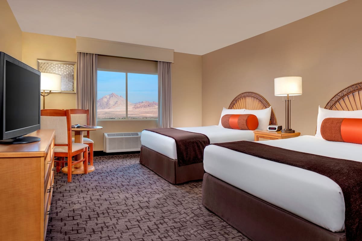 A hotel room with two beds, a desk with a TV, a chair, a lamp, and a view of mountains.