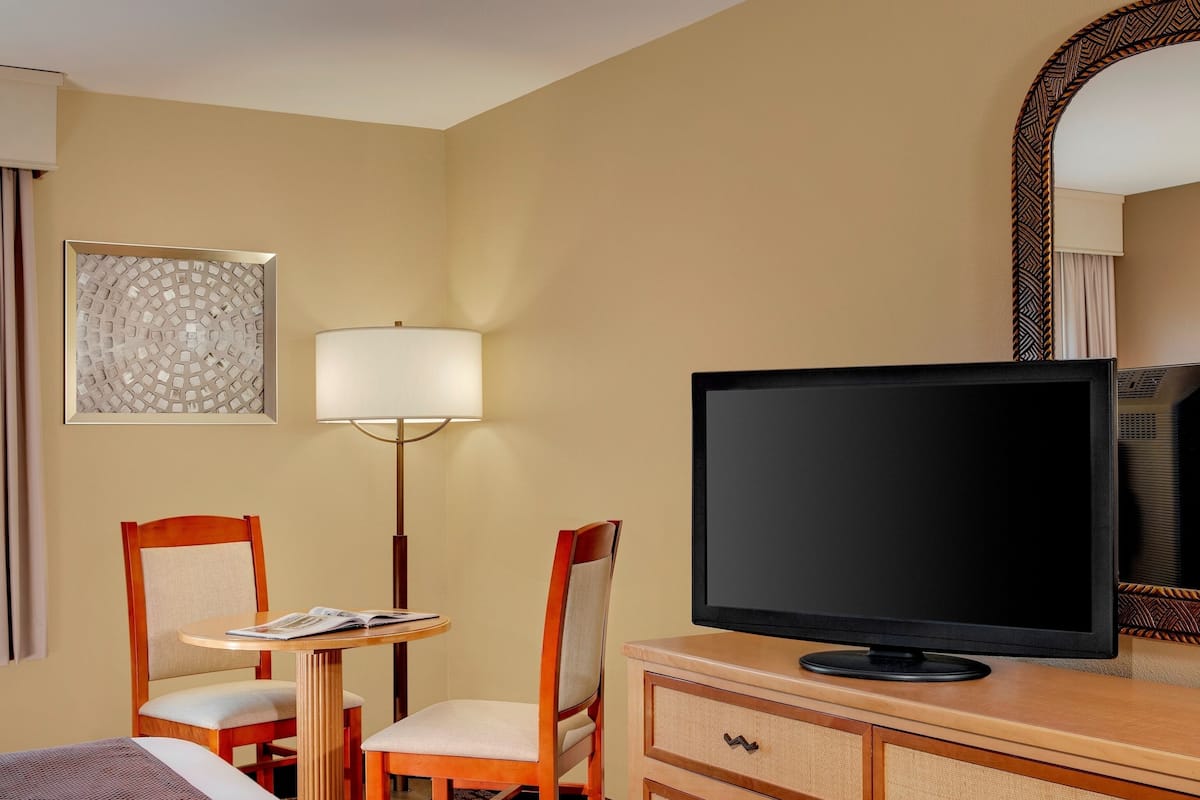 A hotel room with a bed, a television on a wooden dresser, a desk with a chair, a lamp, and a wall-mounted artwork.