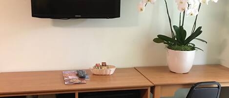 Camera d'albergo con una scrivania in legno, una televisione a parete, un forno a microonde, un piccolo frigorifero, una sedia e una pianta di orchidea in vaso.