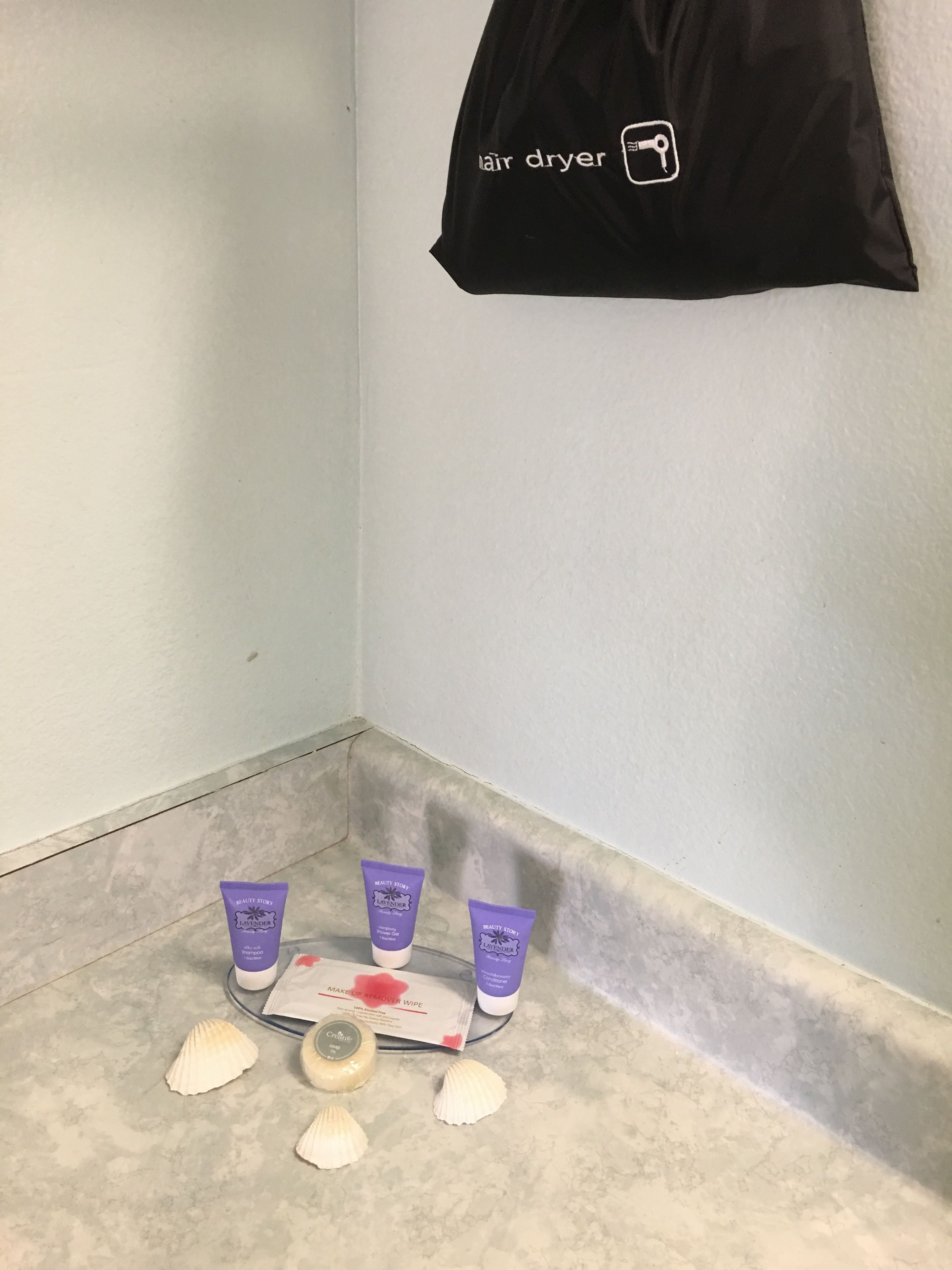 A bathroom corner with a black bag labeled "hair dryer" hanging on the wall, a white towel, and three purple tubes of lotion on a tray.
