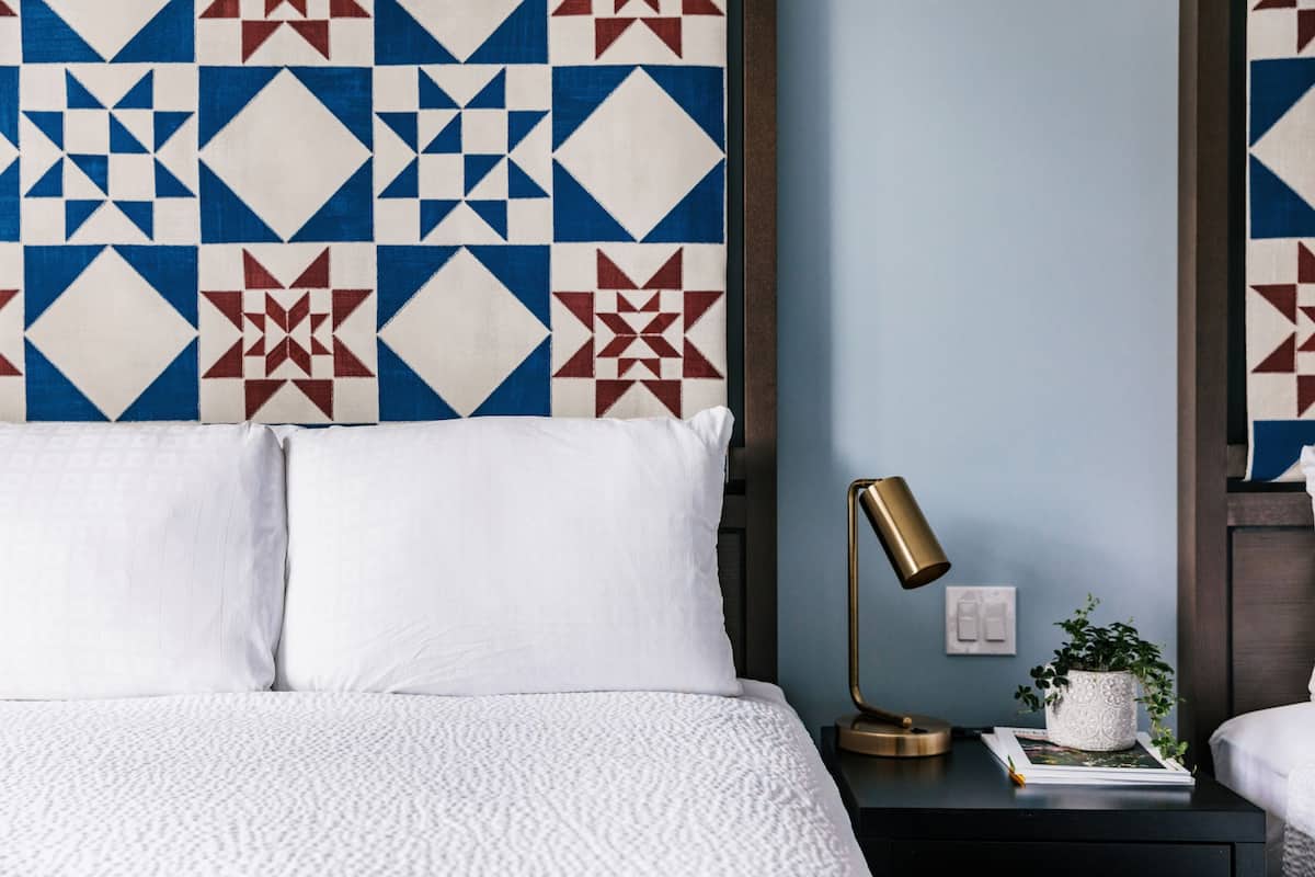 A neatly made bed with white linens, a bedside lamp, a small potted plant, and a wall with a geometric patterned tapestry.