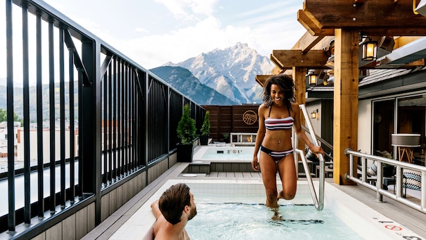 Un couple dans un bain à remous avec vue sur les montagnes, une terrasse en bois et un espace salon extérieur.