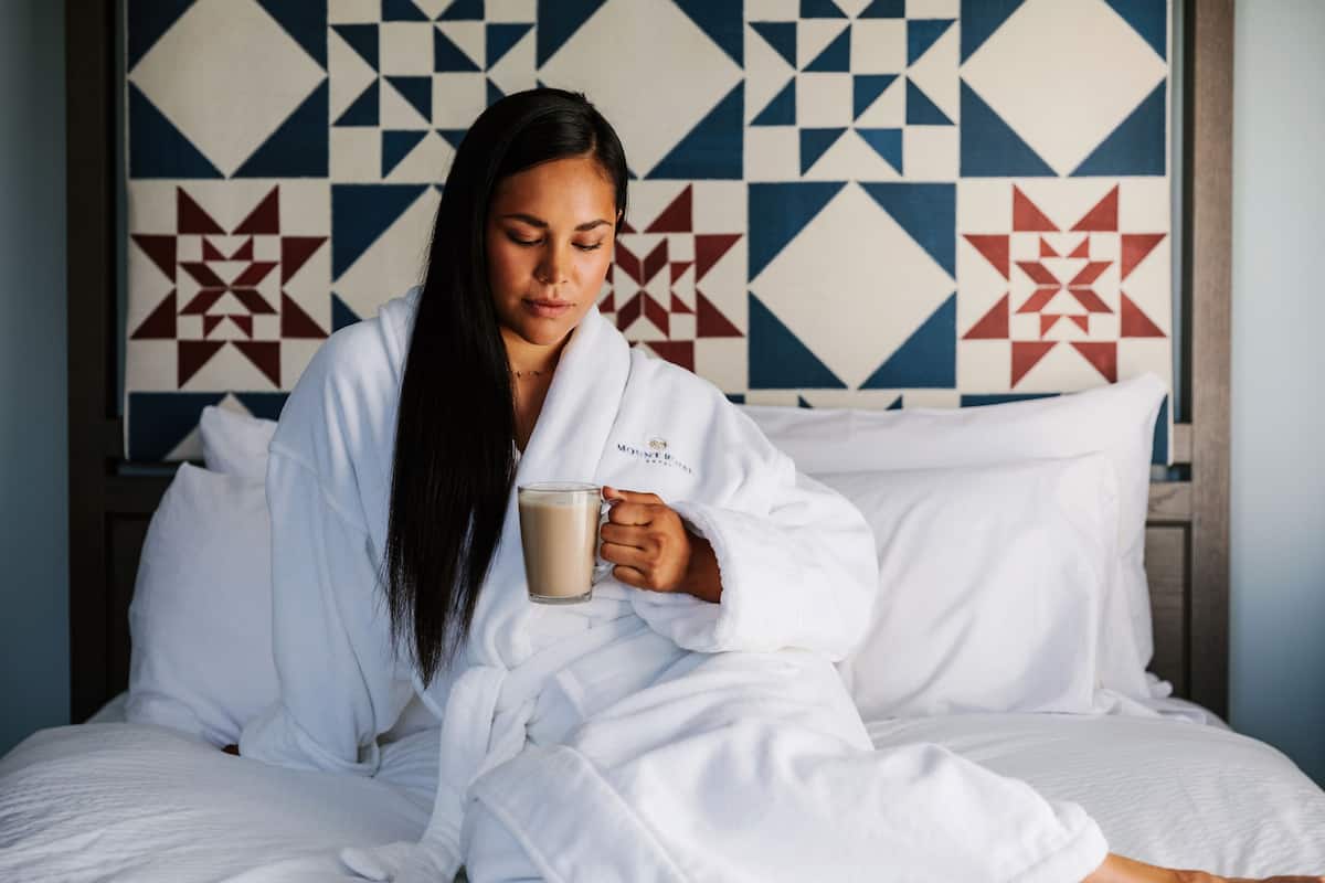 A person in a white robe sitting on a bed with a patterned wall behind them.