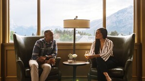 Deux personnes sont assises dans des fauteuils en cuir, face à face, avec une table entre elles. Une lampe est posée sur la table, et de grandes fenêtres offrent une vue sur les montagnes.