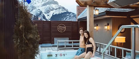 Una pareja junto a una piscina con montañas al fondo, terraza de madera y una bandera.