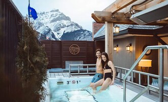 Una pareja junto a una piscina con montañas al fondo, terraza de madera y una bandera.