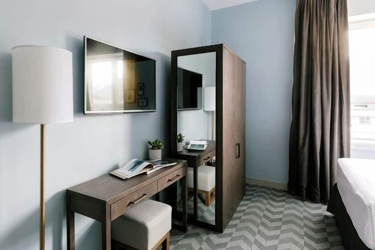 A hotel room with a desk, chair, bedside table, lamp, mirror, wardrobe, and a bed.