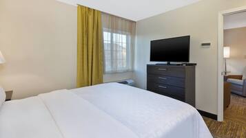 A hotel room with a bed, a television on a dresser, a window with curtains, and a door leading to another room.