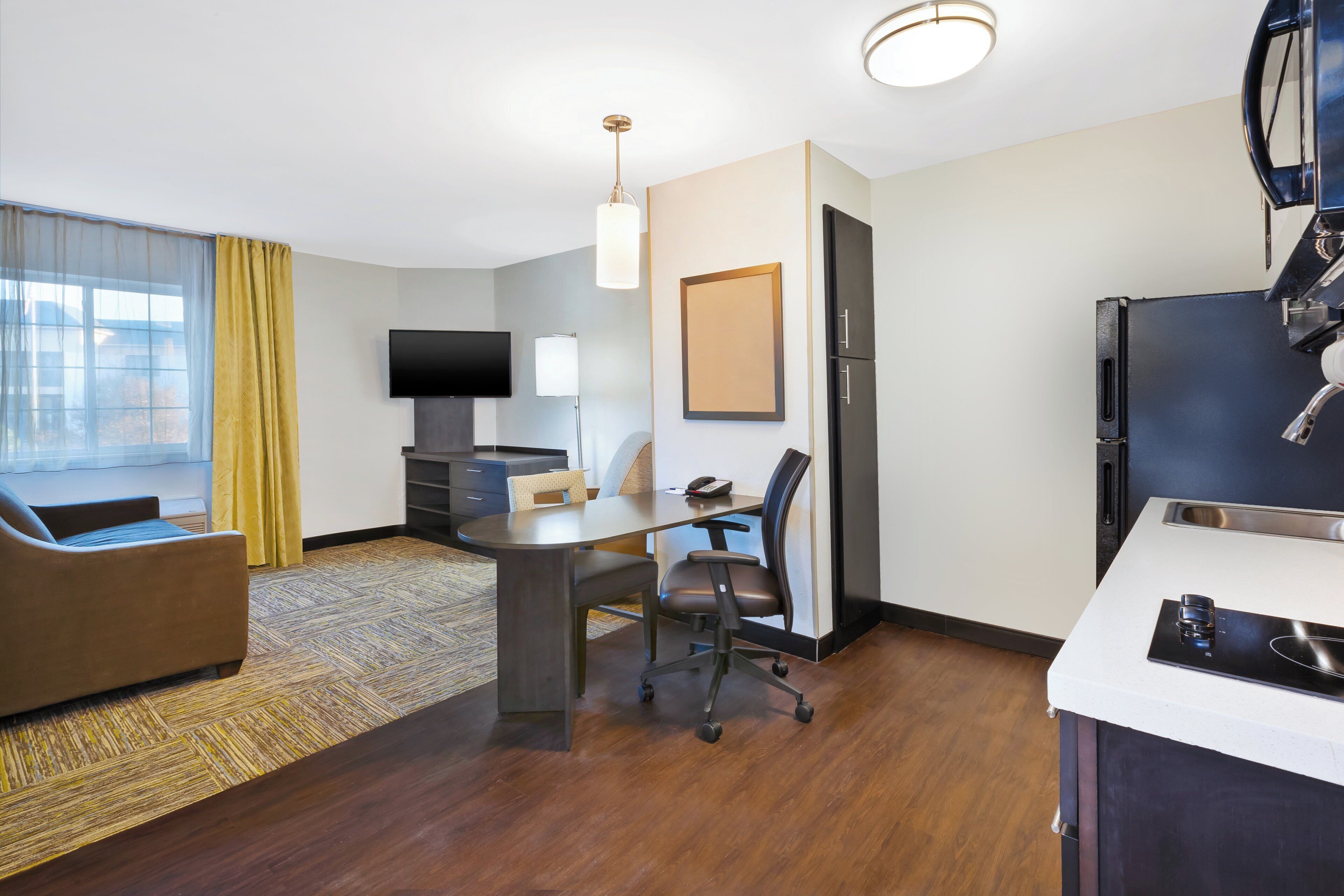 A compact hotel room with a sofa, a dining table, a TV, a desk, and a kitchenette.