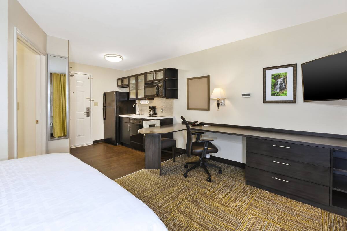 A hotel room with a bed, a desk, a chair, a refrigerator, a microwave, a sink, and a TV.