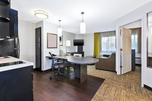 A modern hotel room with a dining area, a flat-screen TV, a sofa, and a large window with curtains.