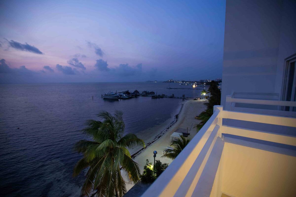 Vista costera desde un balcón con vista a una playa, barcos amarrados, palmeras y un horizonte urbano distante.