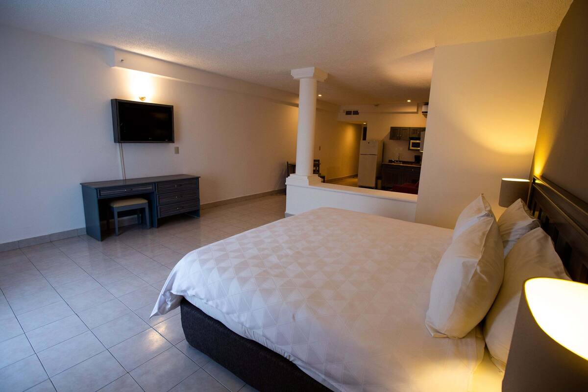 Habitación de hotel con cama, televisor montado en la pared, escritorio y una cocina al fondo.
