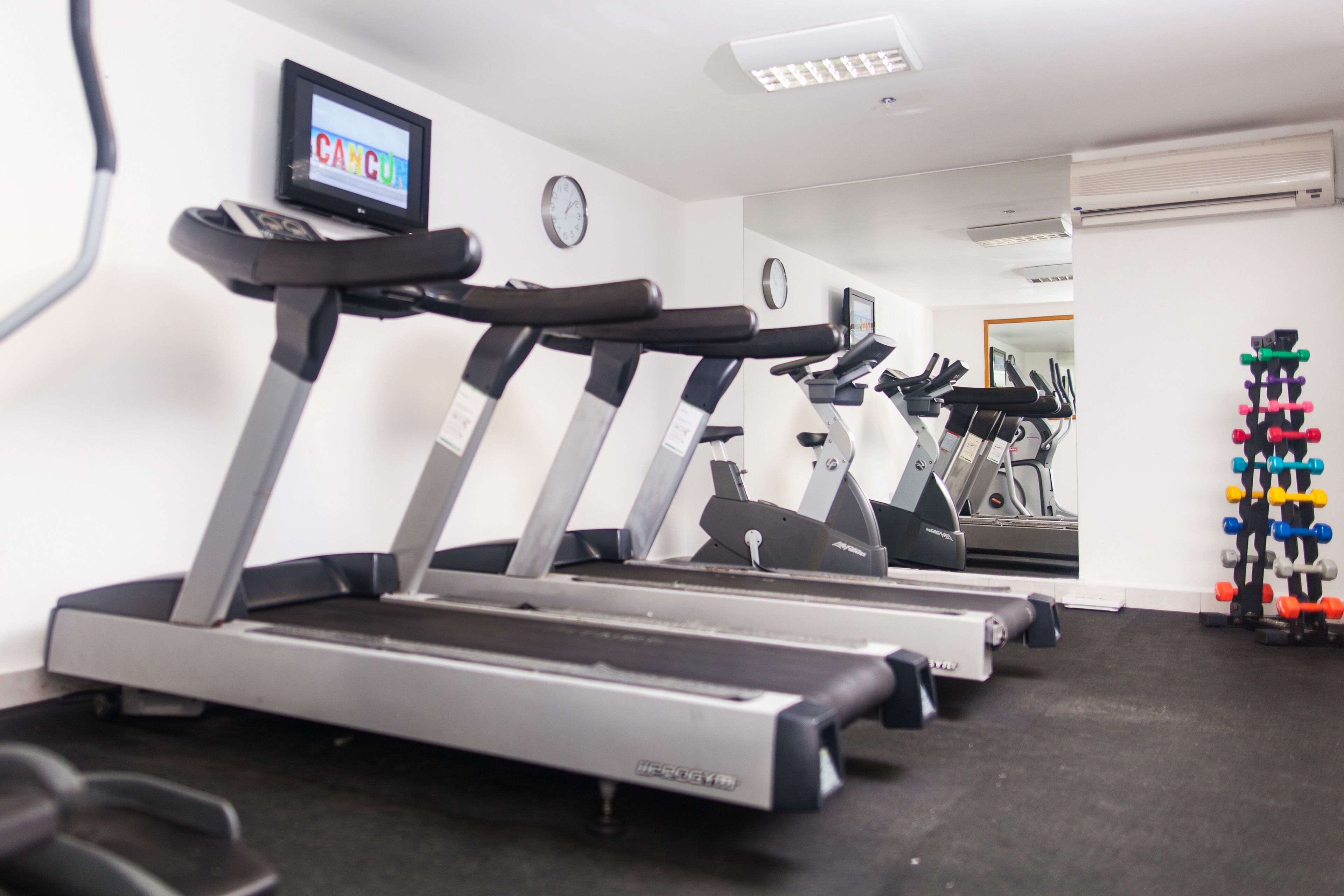 A gym with multiple treadmills, a wall-mounted TV displaying 'CANCUN', and a mirror reflecting the room.