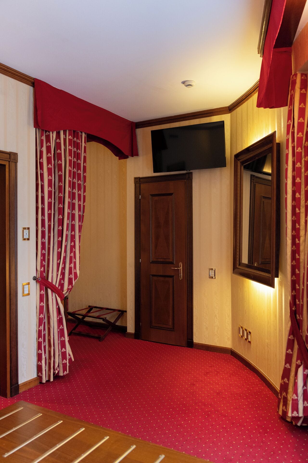 Quarto de hotel com carpete vermelho, porta de madeira, televisão de tela plana acima de uma porta de madeira e cortina vermelha com padrão floral.