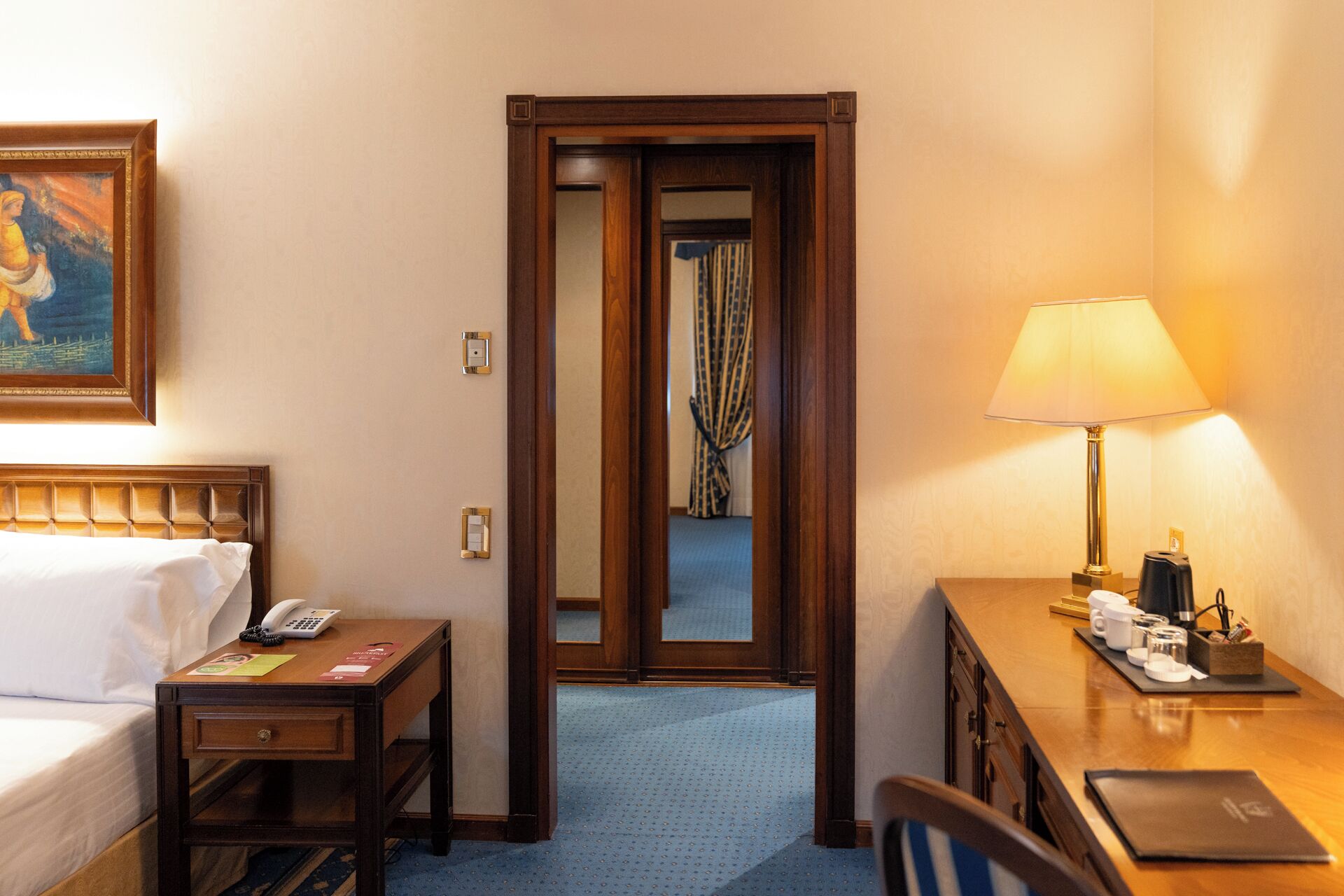 A hotel room with a bed, bedside table with a lamp, a desk with a laptop, and a mirror.