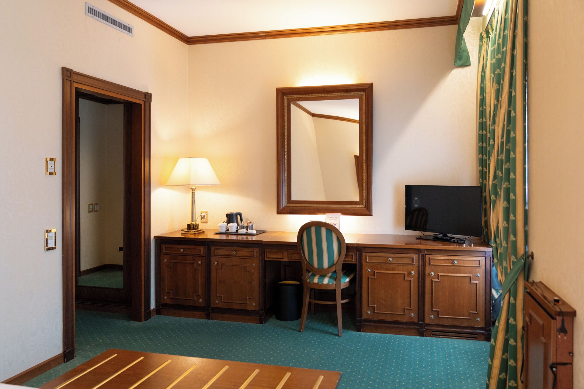 A hotel room with a wooden desk, a chair, a television, a mirror, and a lamp.