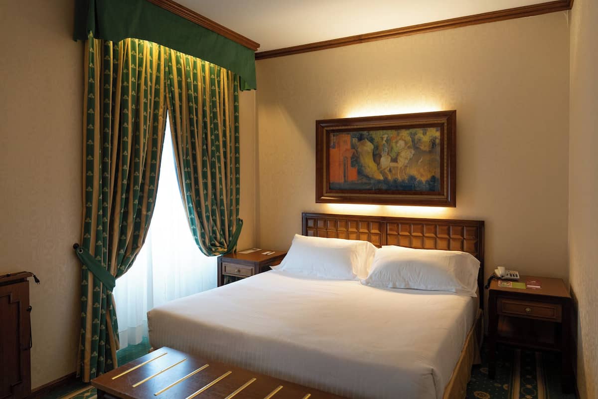Una habitación de hotel con una cama, una mesita de noche, un cuadro en la pared y una ventana con cortinas.