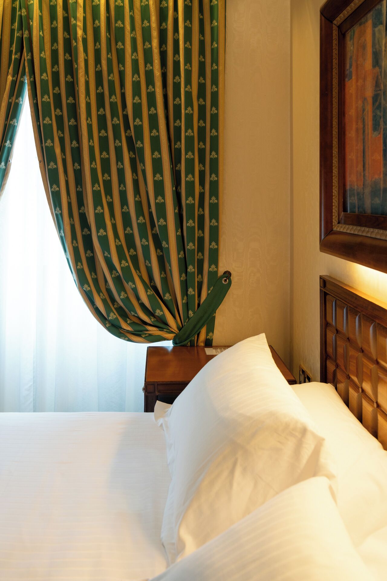 Una habitación de hotel con cama, mesita de noche, un cuadro en la pared y cortinas.