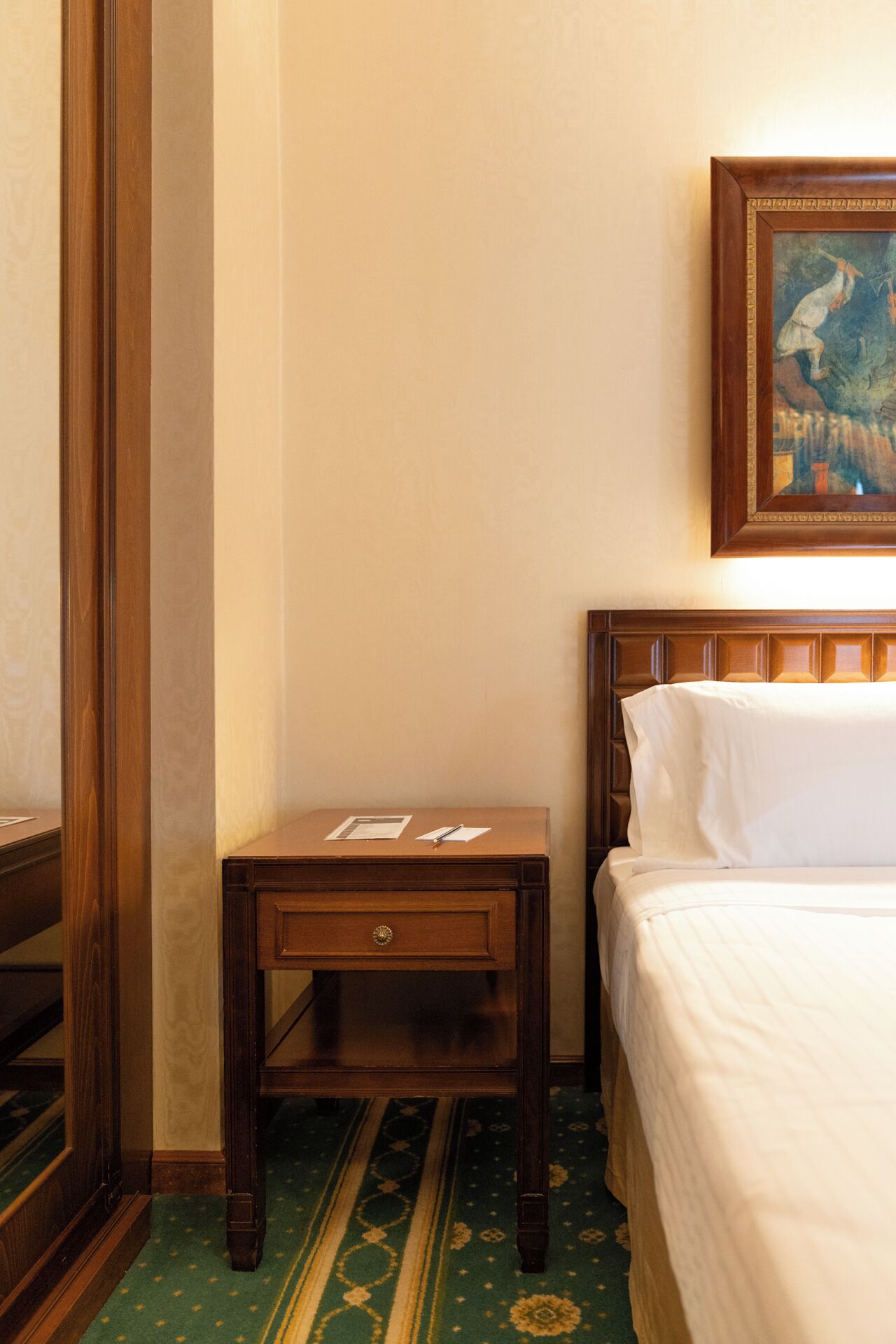 A hotel room with a wooden nightstand, a bed with white linens, and a framed painting on the wall.