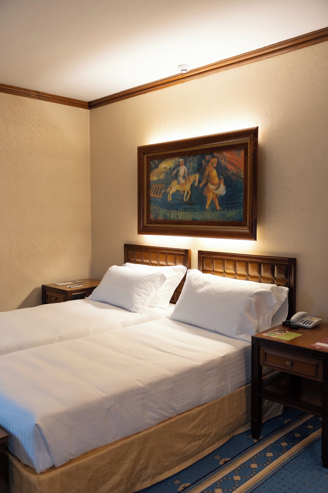 A hotel room with a large bed, wooden headboard, bedside tables, a painting on the wall, and a blue patterned rug.