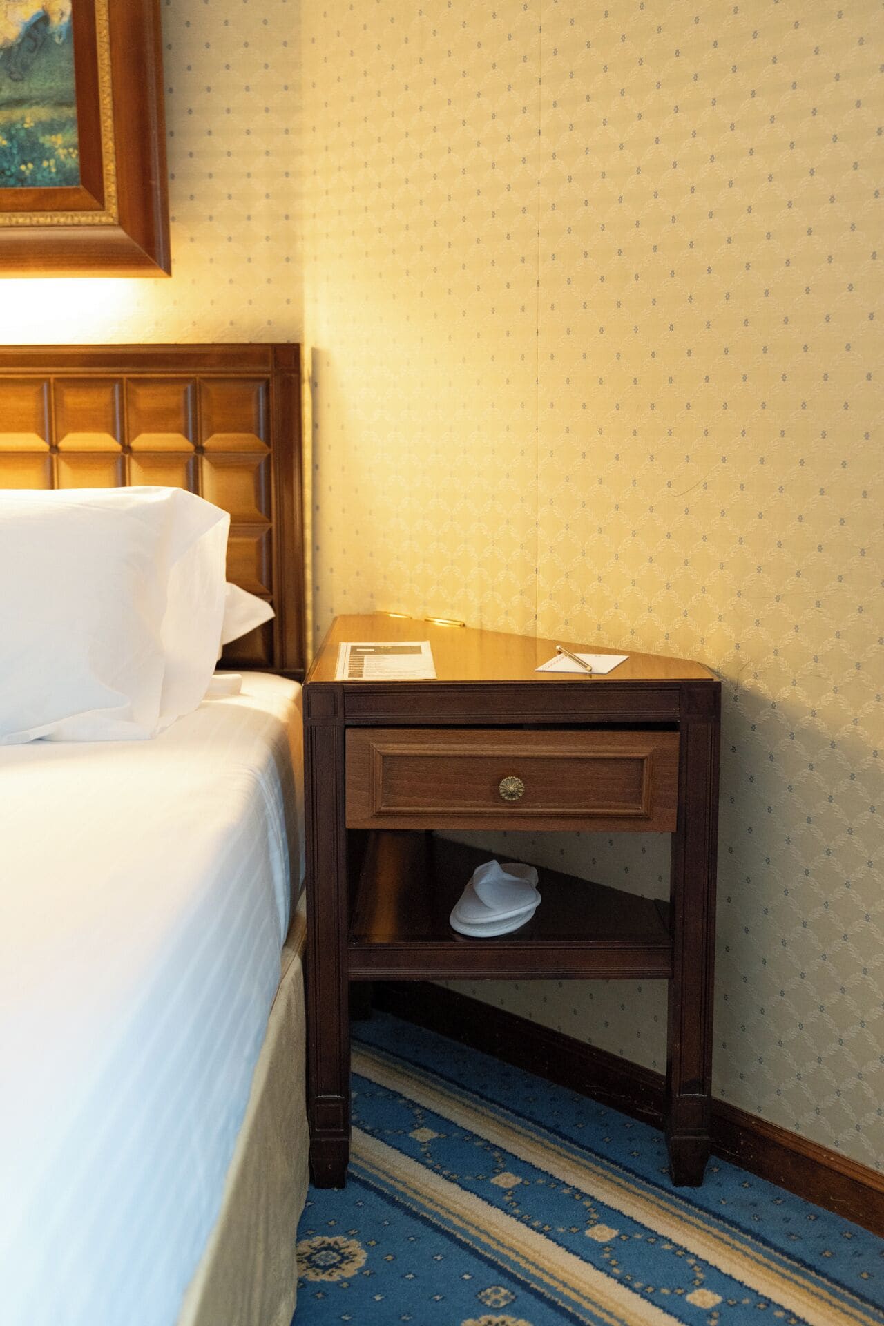 Una habitación de hotel con una cama bien hecha, una mesita de noche de madera con cajón y un papel tapiz estampado.