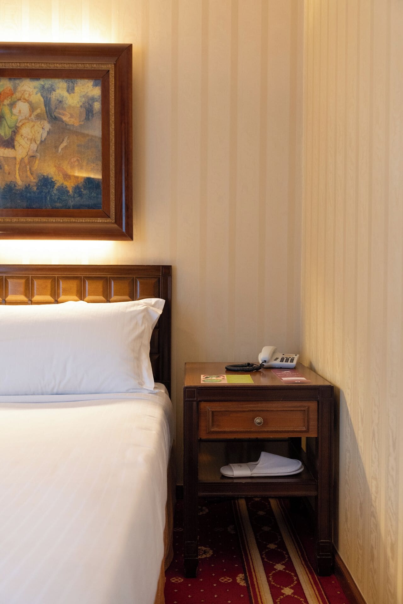 A hotel room with a bed, a nightstand with a telephone and a notepad, and a painting above the bed.