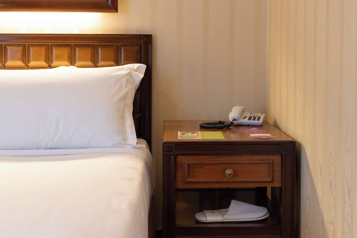 Una habitación de hotel con una cama, una mesita de noche con un teléfono y un bloc de notas, y un cuadro sobre la cama.