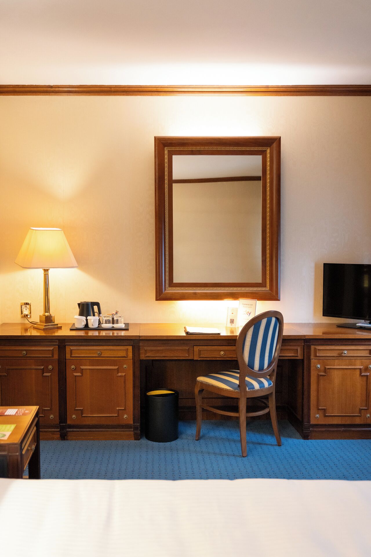 A hotel room with a bed, a desk with a lamp, a chair, a television, and a trash can.
