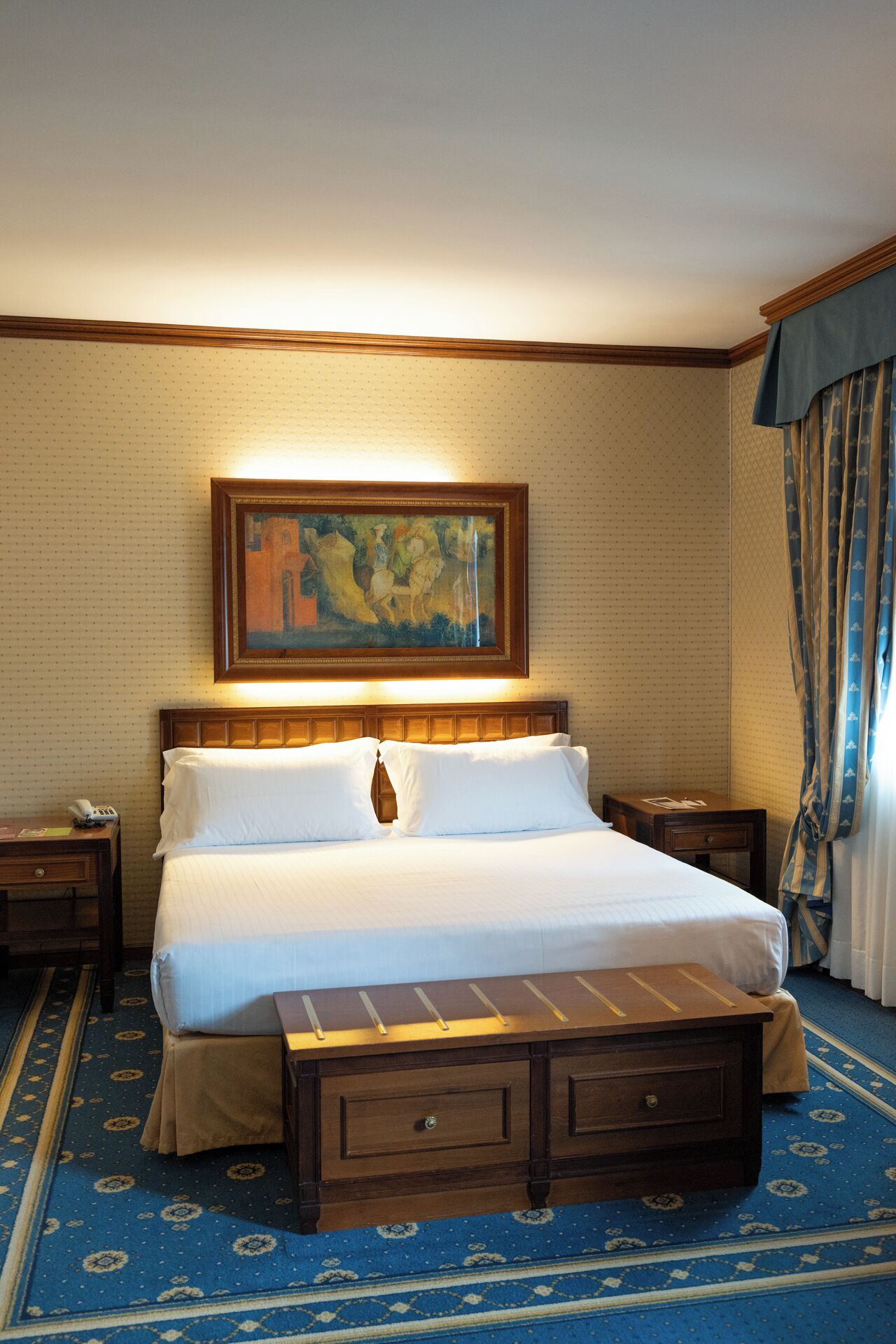 Une chambre d’hôtel avec un grand lit, une table de chevet, une lampe de chevet, un tableau au-dessus du lit et une moquette à motifs.