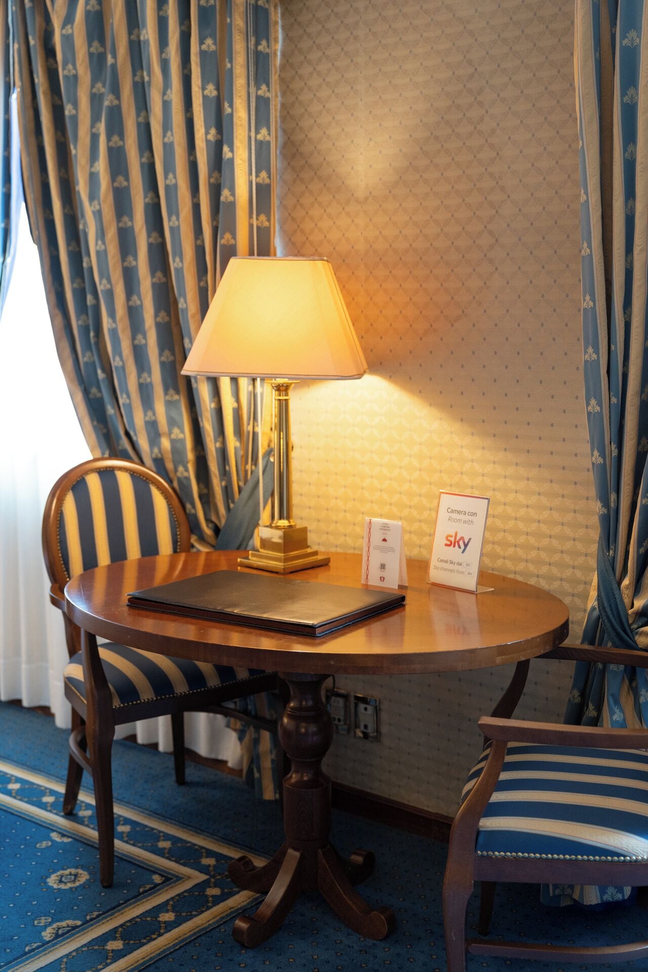 Ein Hotelzimmer mit einem runden Holztisch, einer Lampe, einem Stuhl, einem Schild, das auf eine Kamera in dem Raum hinweist, und einer Wand mit Muster.