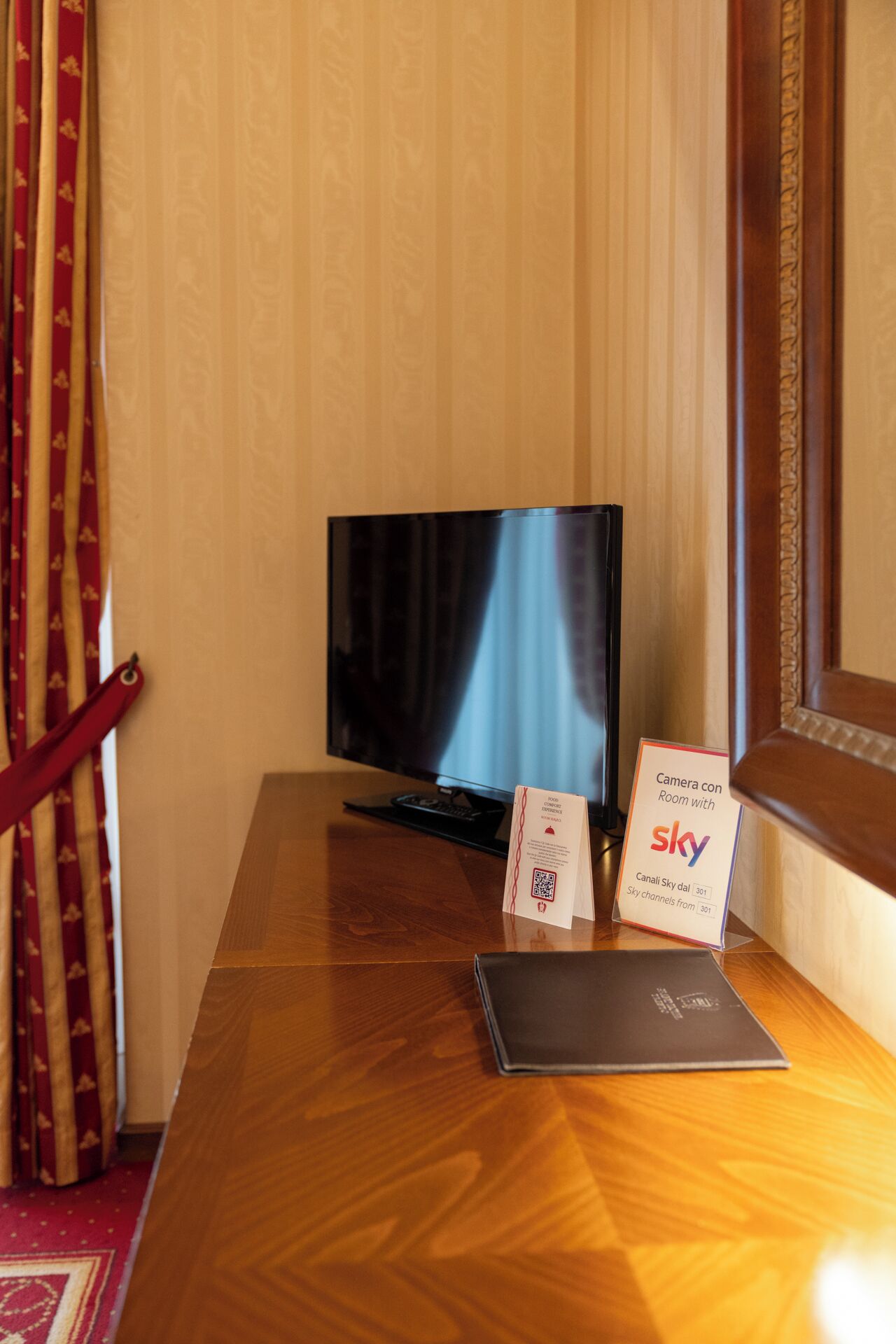 Une chambre d’hôtel avec un bureau en bois, une télévision, un panneau indiquant la chaîne Sky, et un rideau rouge.