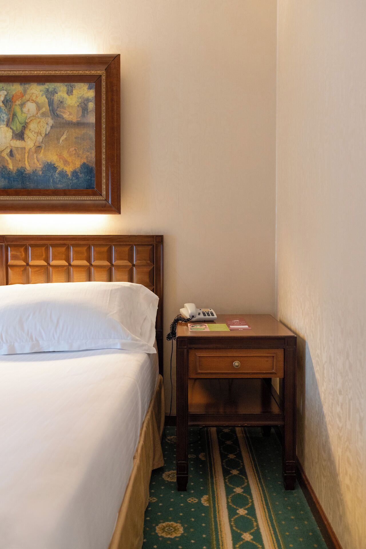 Une chambre d’hôtel avec un lit, une table de chevet en bois avec un téléphone et un tableau au-dessus du lit.
