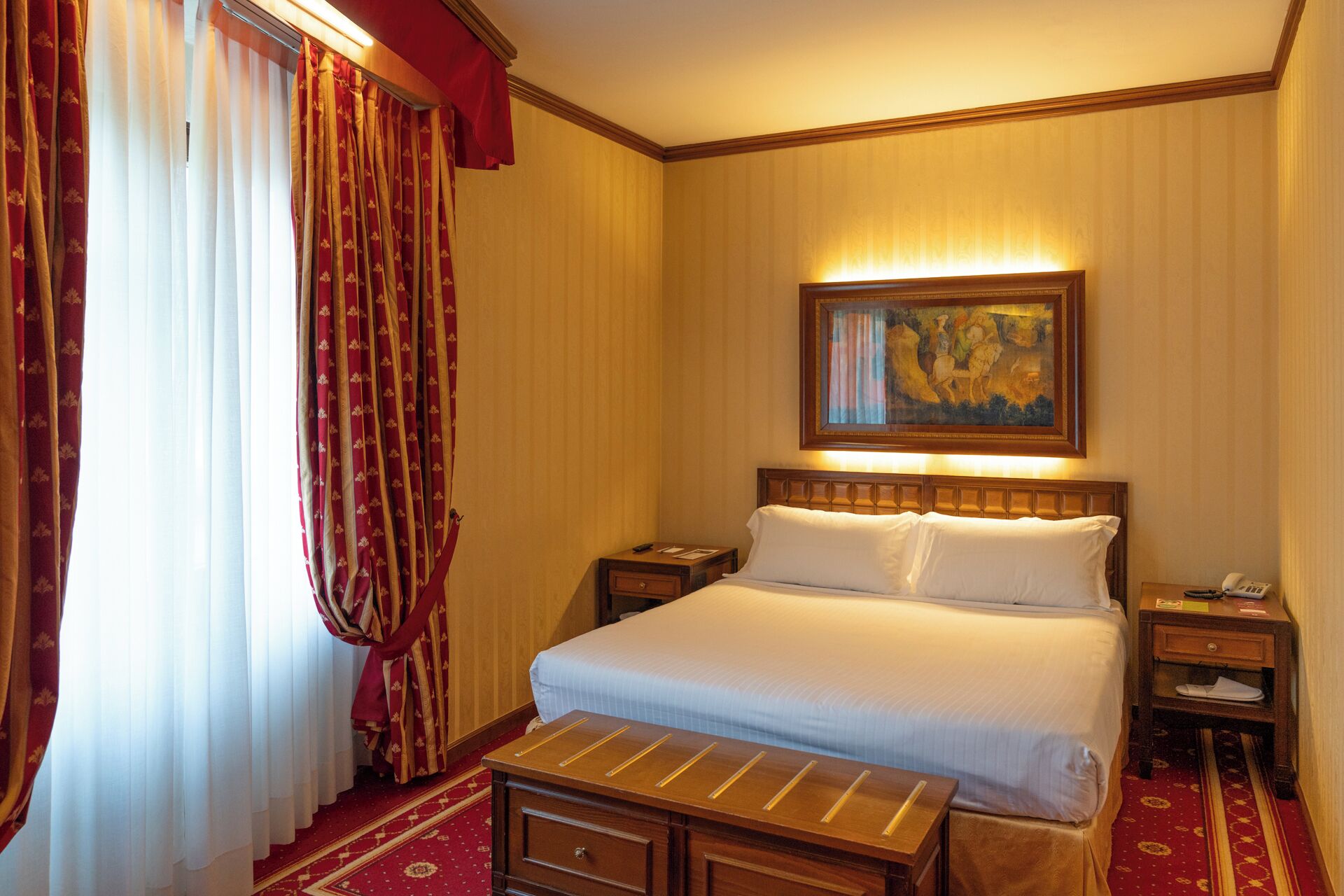 Habitación de hotel con una cama grande, una mesita de noche, un cuadro en la pared y una ventana con cortinas.